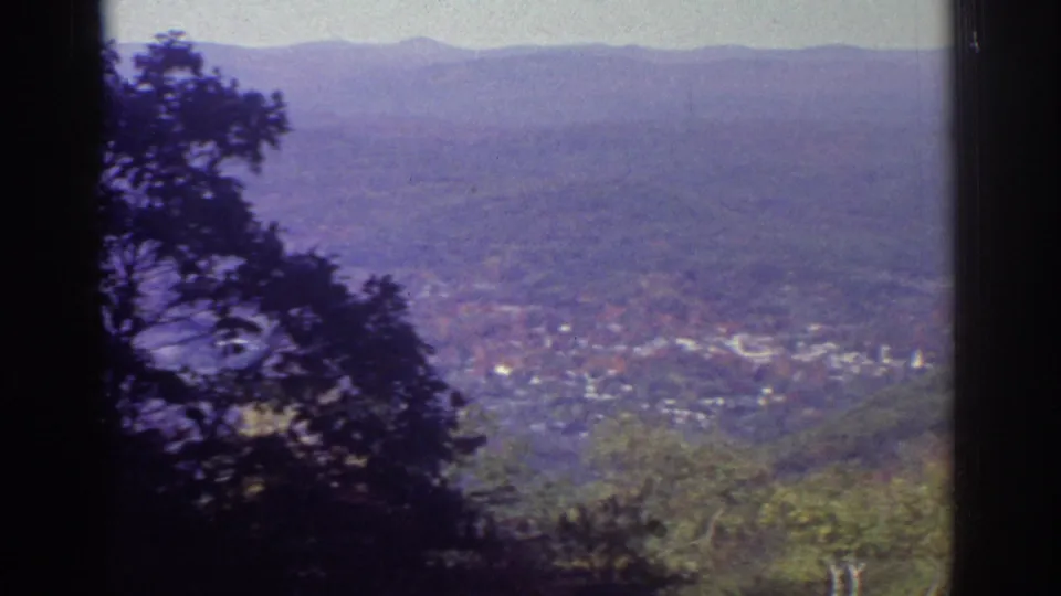 1976 Watkins Glen valley panorama showing distant village buildings, wooded foreground, hazy hills, and soft blue-purple 8mm color.