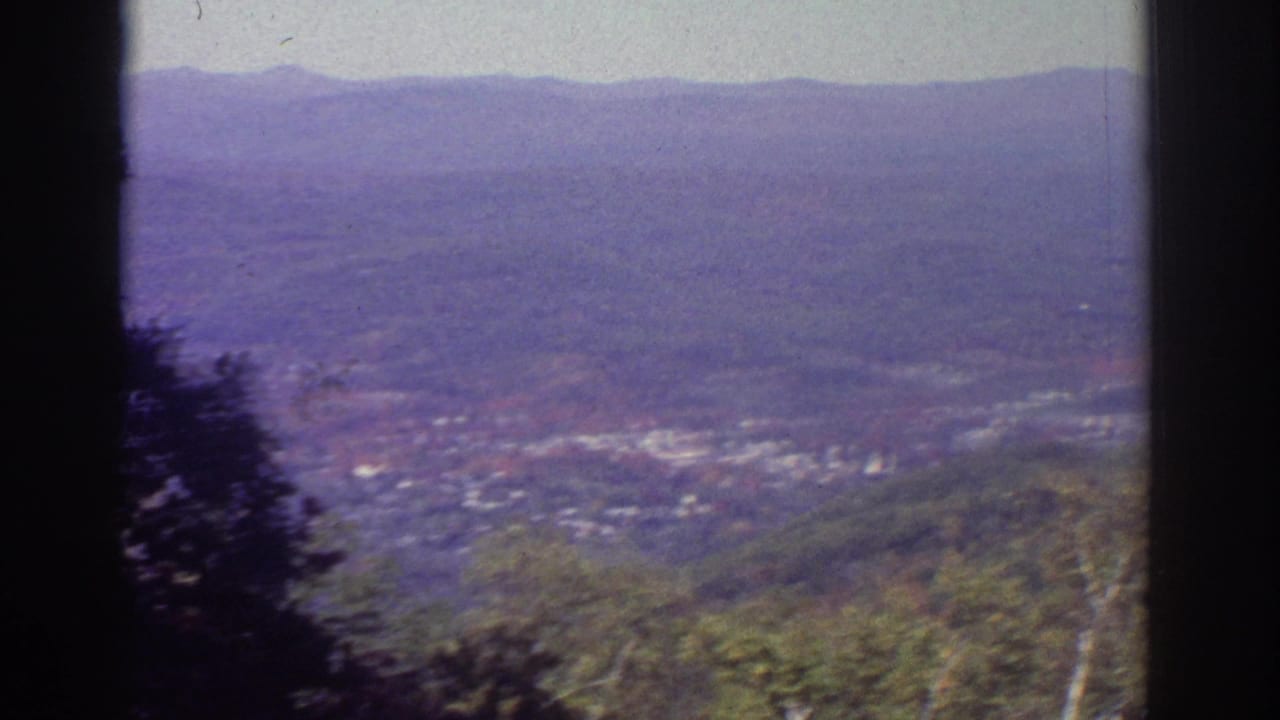 1976 Watkins Glen-area home-movie view of a hazy valley town, wooded foreground, and distant mountain ridge framed by dark window edges.