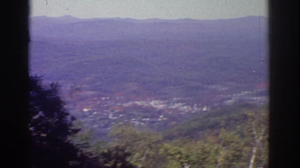 1976 Watkins Glen-area home-movie view of a hazy valley town, wooded foreground, and distant mountain ridge framed by dark window edges.