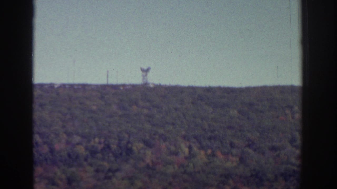 1976 Watkins Glen home-movie frame showing a wooded autumn hillside, pale sky, film edges, and a small distant ridge structure.