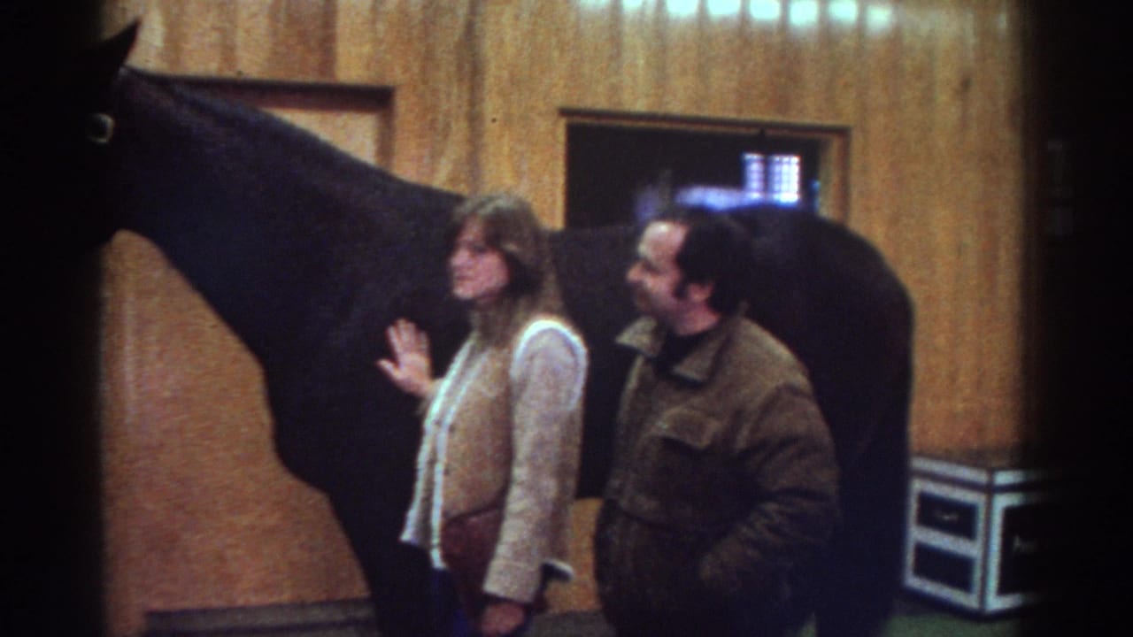 Watkins Glen, New York home-movie frame from 1976 showing adults near a dark horse inside a wood-paneled stable.