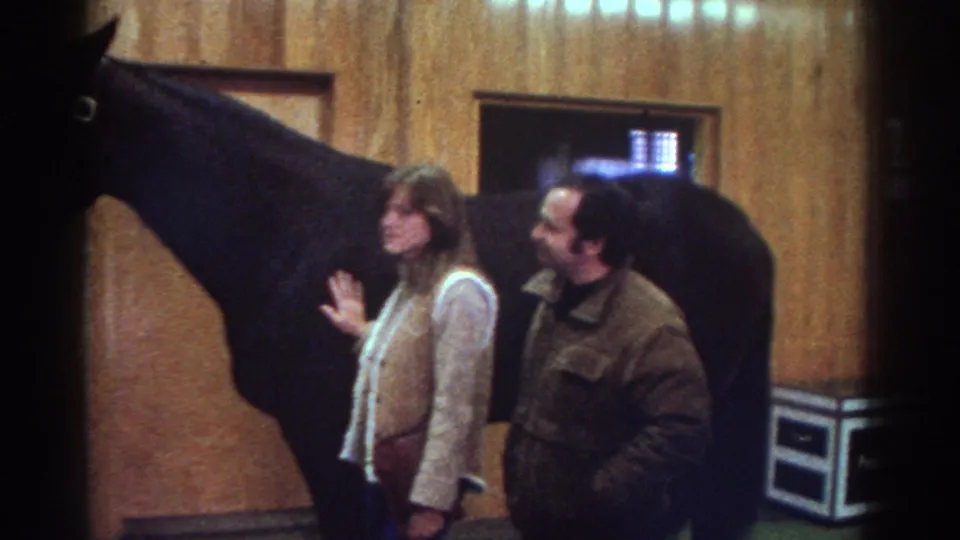 Watkins Glen, New York home-movie frame from 1976 showing adults near a dark horse inside a wood-paneled stable.