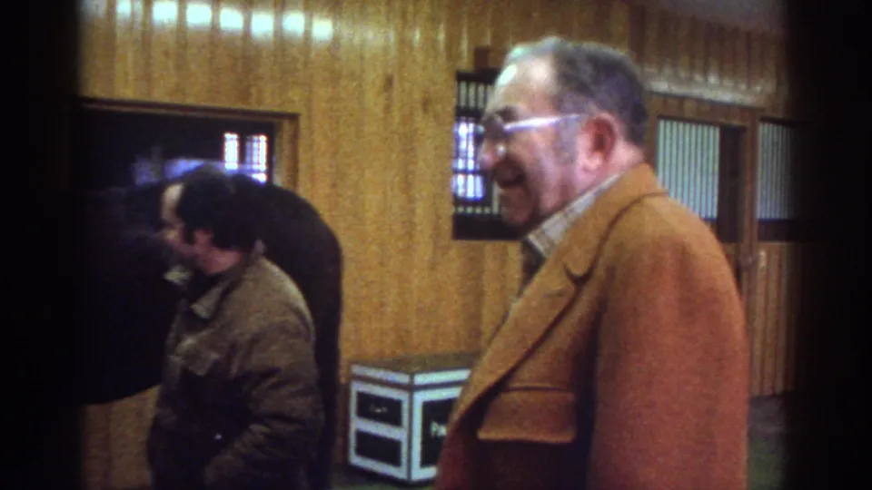 Watkins Glen stable frame showing two adult men near a dark horse, with wood walls and stall details visible.