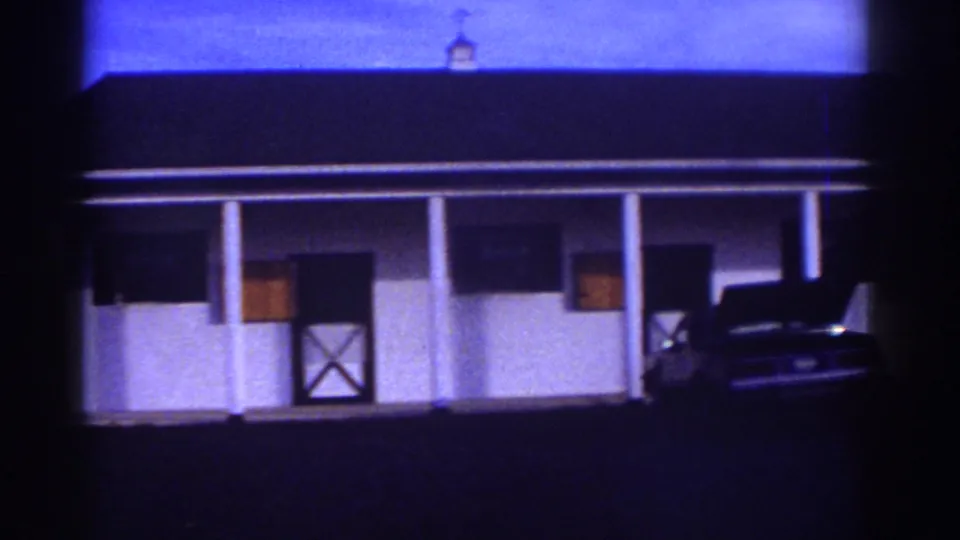 Exterior view of a white stable building at dusk in the Watkins Glen home movie, with a parked car nearby.