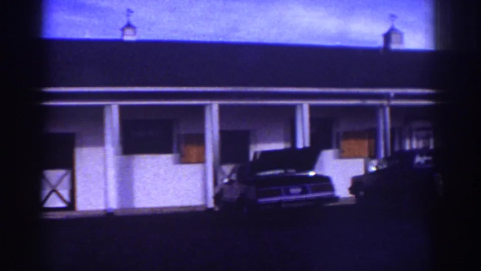 1976 Watkins Glen home-movie frame showing a white motel-style exterior, porch columns, and parked cars under a dark roof.