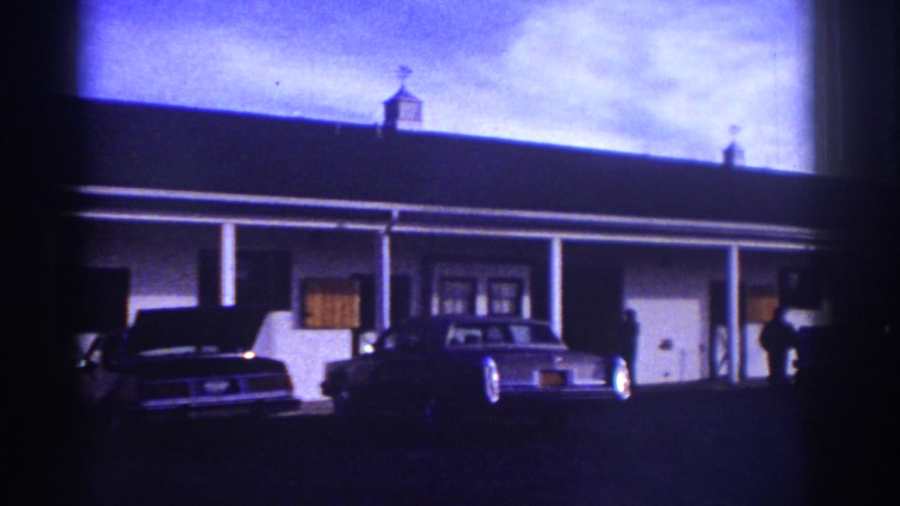 Watkins Glen 1976 frame with a motel-style building, porch, classic sedan, and a person standing near the right side.