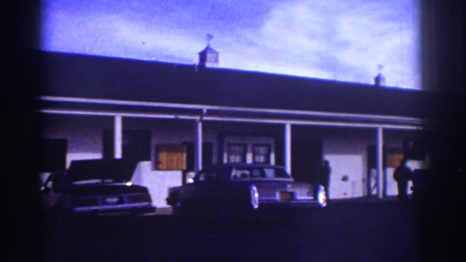 Watkins Glen 1976 frame with a motel-style building, porch, classic sedan, and a person standing near the right side.