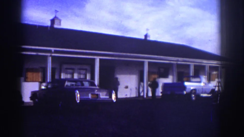 1976 Watkins Glen frame showing a white motel-style exterior with a sedan, blue pickup truck, people, and a ladder near the truck.