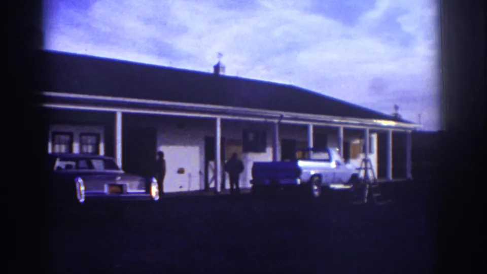 Watkins Glen 1976 home-movie frame with a motel-style porch, blue pickup, classic sedan, standing people, and ladder.