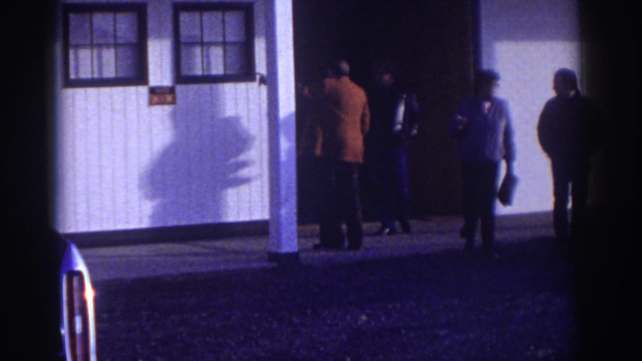 In Watkins Glen, New York, 1976, four adult men appear outside a white building with a porch, windows, and a dark doorway.