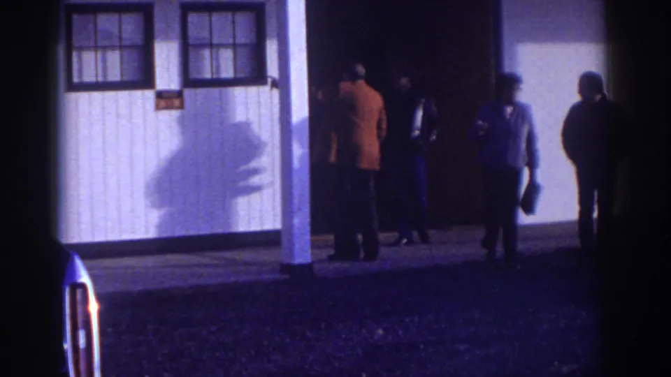 In Watkins Glen, New York, 1976, four adult men appear outside a white building with a porch, windows, and a dark doorway.