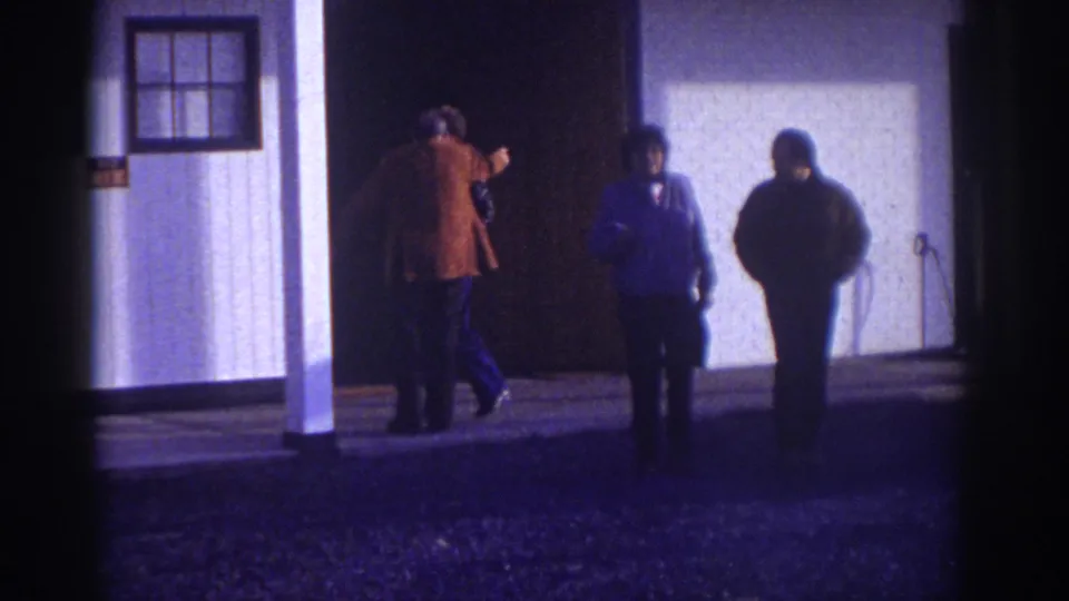 Several adult men are seen outside the Watkins Glen building in 1976, with one man in a brown jacket near the entrance.