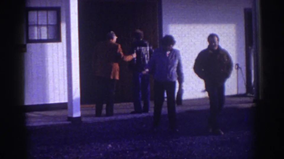 The 1976 Watkins Glen home-movie frame shows four adult men outside a white building, with two closer to the camera and two near the doorway.