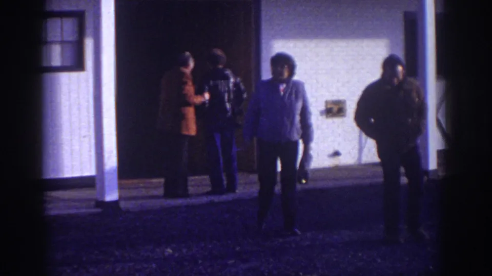At the end of the Watkins Glen clip, adult men remain outside the white building while two figures move toward the camera.