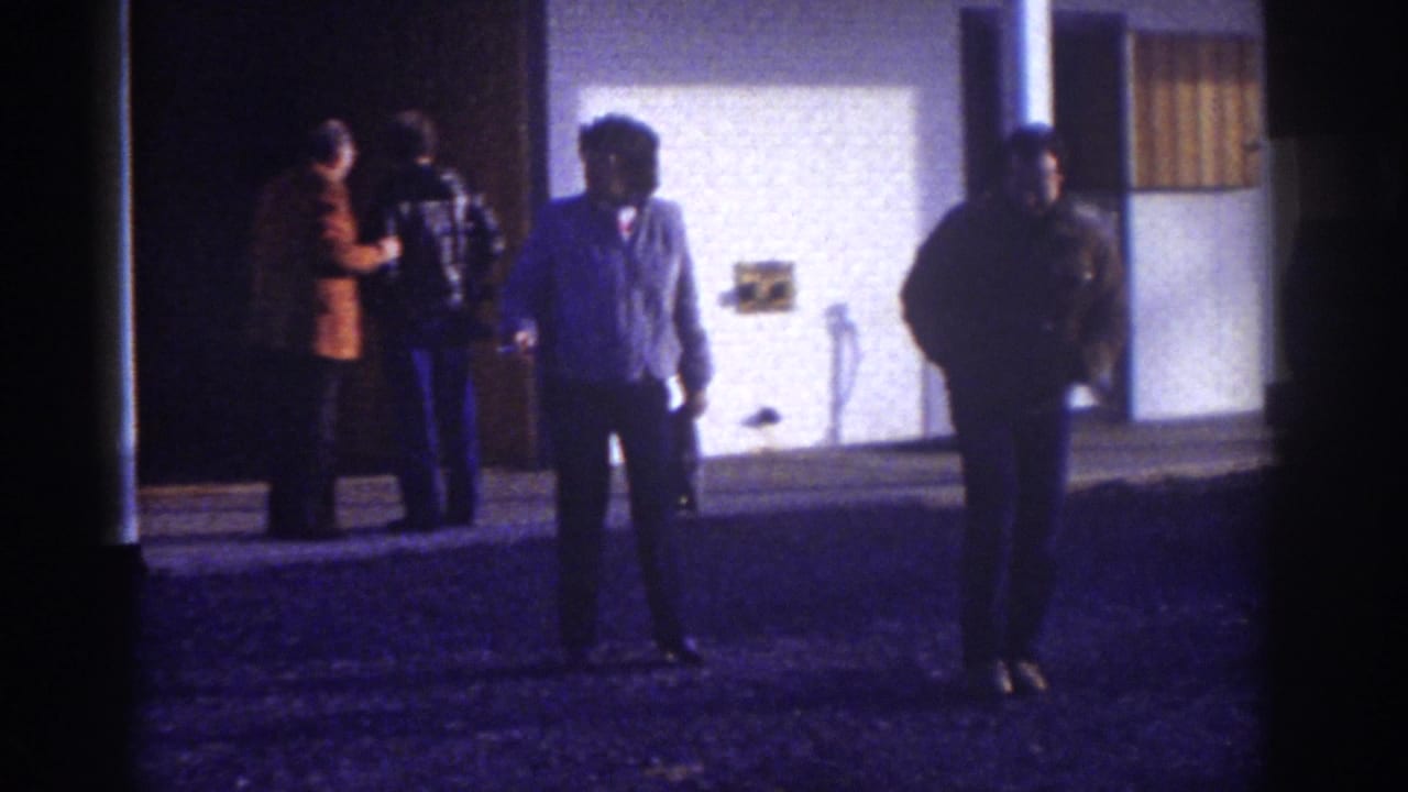 1976 Watkins Glen home-movie frame showing bundled adults outside a motel at night, partly silhouetted by a lit exterior wall.