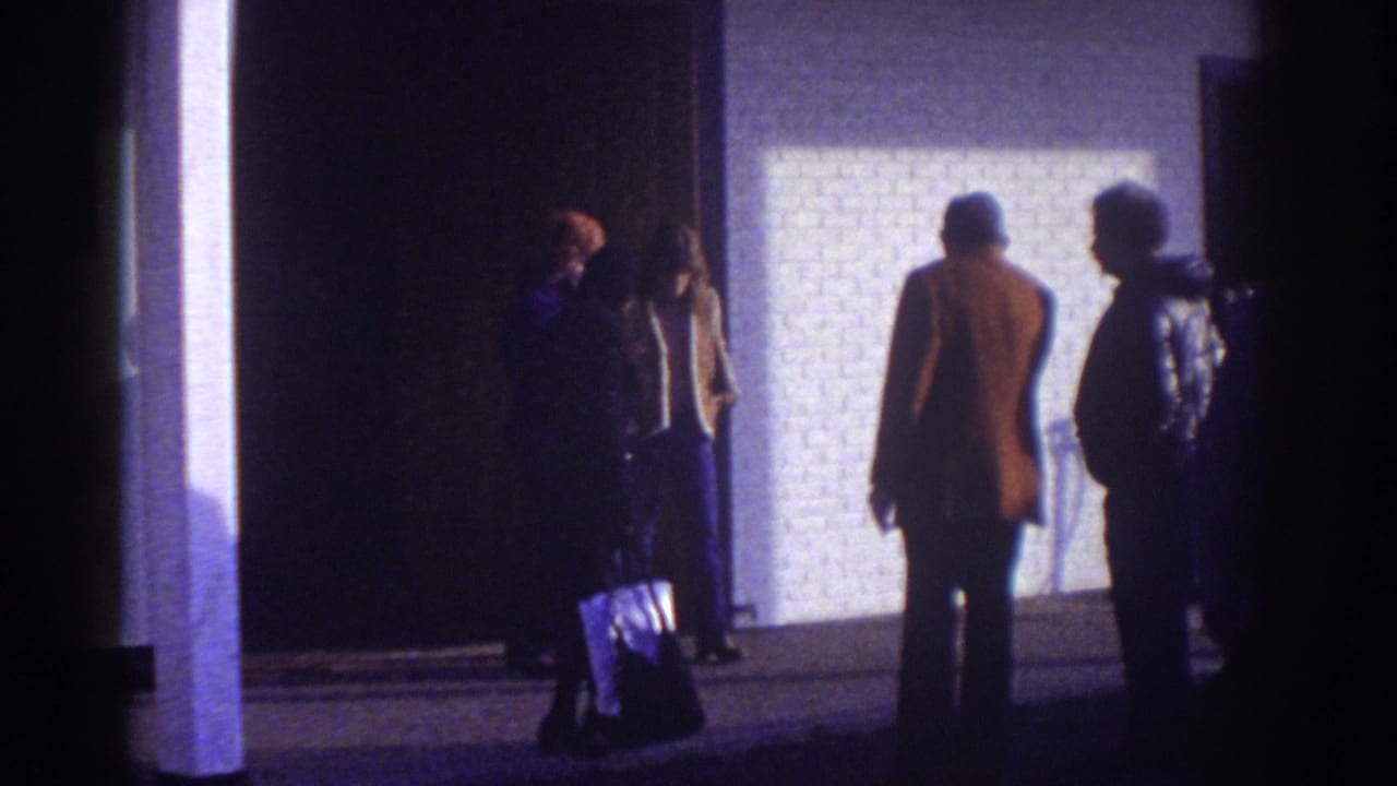 Watkins Glen motel exterior at night with adults in coats standing near a lit wall and dark entrance, one holding a bag.