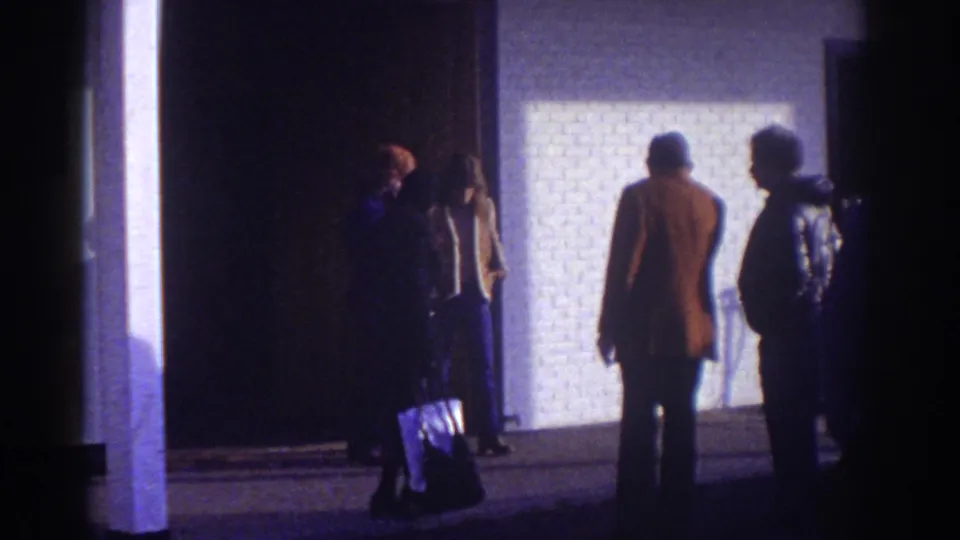 Watkins Glen motel exterior at night with adults in coats standing near a lit wall and dark entrance, one holding a bag.