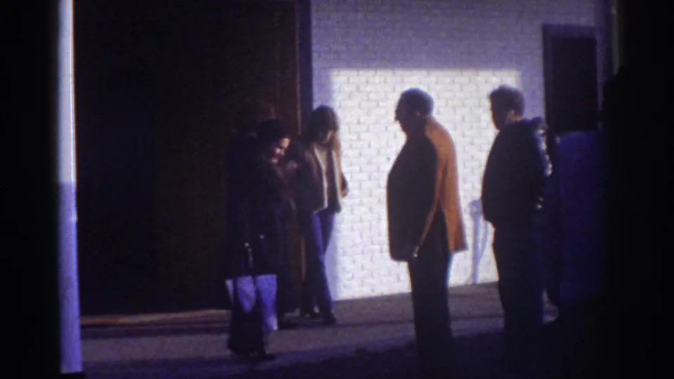 Nighttime 8mm frame of adults in winter coats outside a Watkins Glen motel, seen as silhouettes against a bright exterior wall.