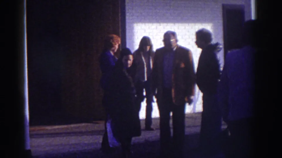 1976 home-movie frame from Watkins Glen showing adults gathered outside a motel doorway, lit from behind by a pale wall.