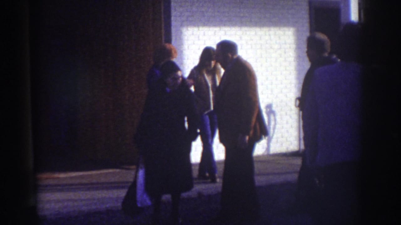 Watkins Glen 1976 home-movie frame showing several people standing near a lit brick building exterior at night.