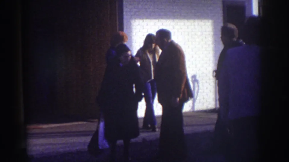 Watkins Glen 1976 home-movie frame showing several people standing near a lit brick building exterior at night.