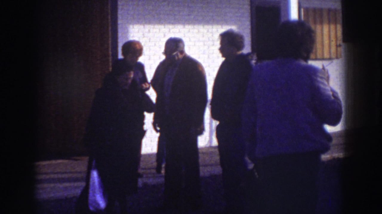 Watkins Glen 1976 frame with adults in coats standing and talking outside a brick building under strong exterior light.