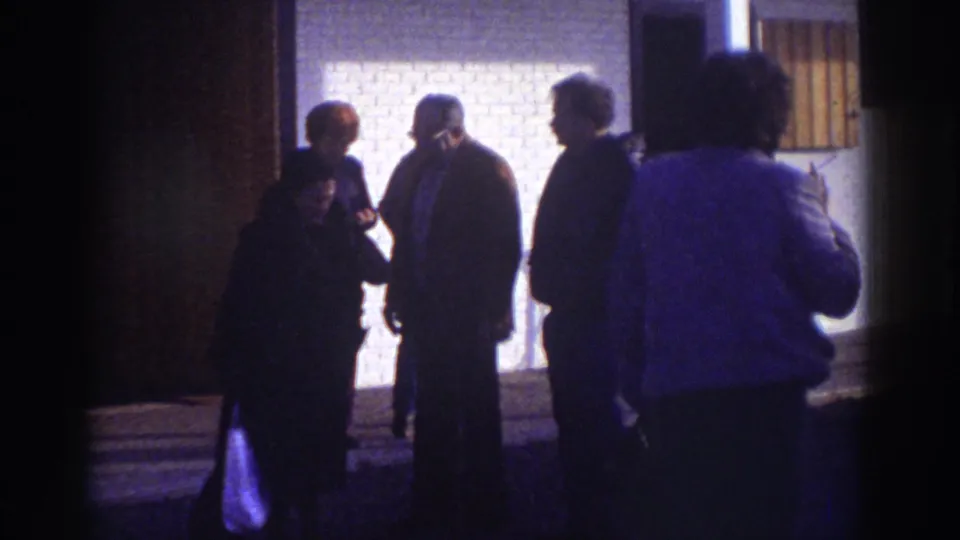 Watkins Glen 1976 frame with adults in coats standing and talking outside a brick building under strong exterior light.