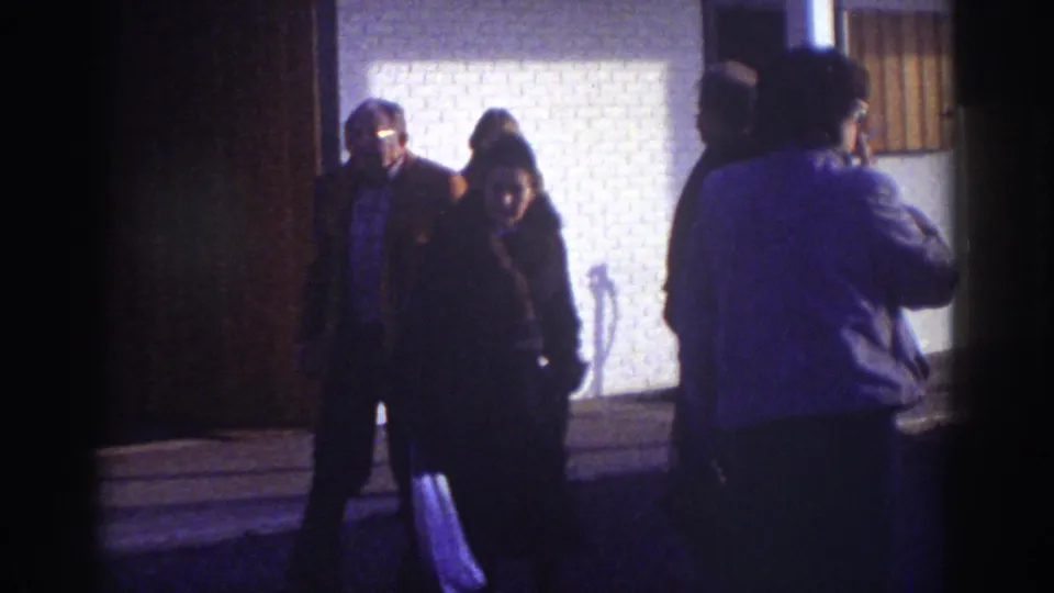 Watkins Glen 1976 frame showing two foreground figures near the camera with other people by a bright brick wall.