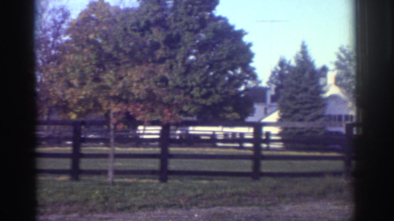 Watkins Glen 1976 home-movie frame showing a fenced lawn, mature trees, and a white house in the distance.