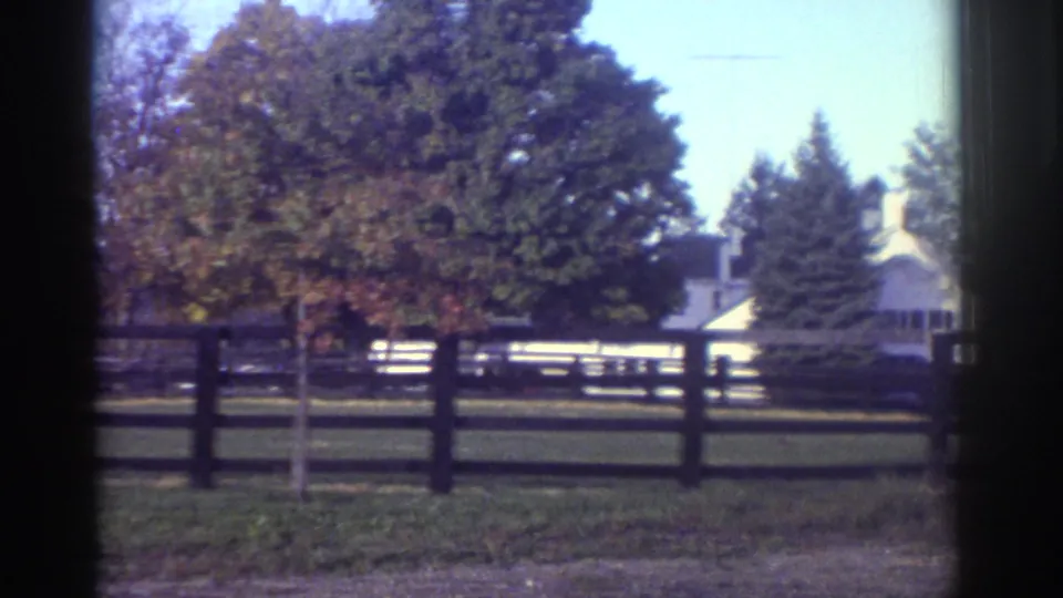 Watkins Glen 1976 home-movie frame showing a fenced lawn, mature trees, and a white house in the distance.