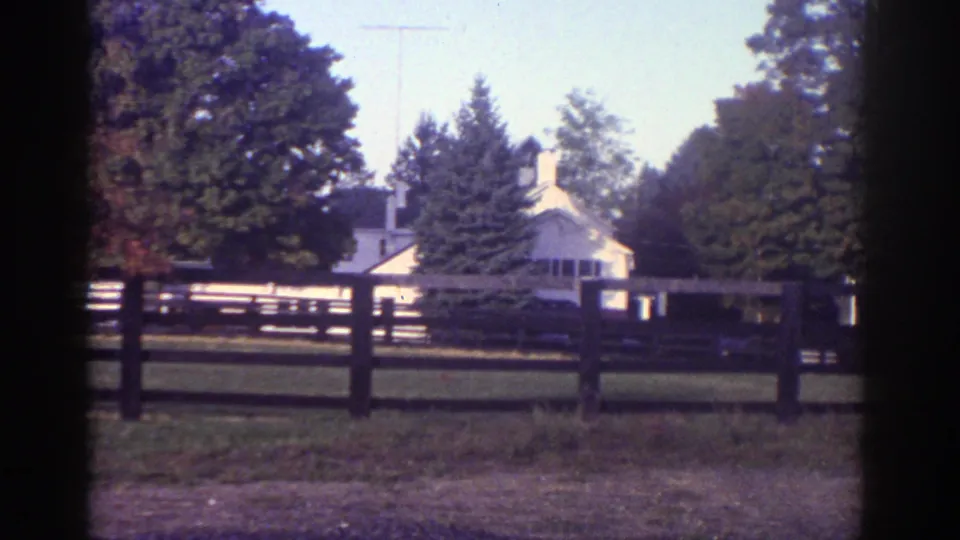Watkins Glen home-movie frame showing the rural house more centrally behind the fence and trees.