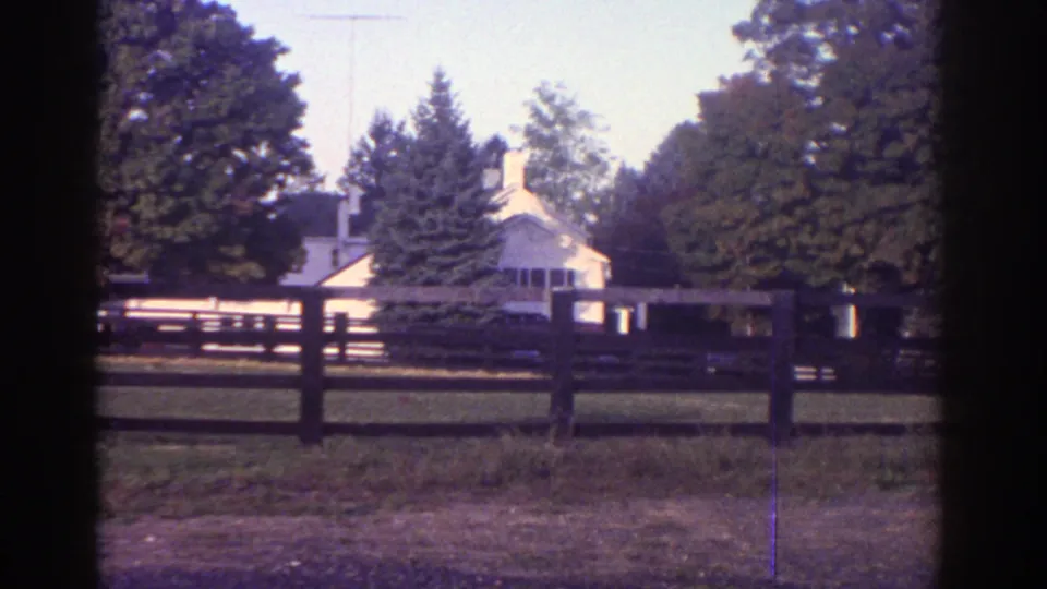 Final Watkins Glen frame with a white house set beyond a rail fence, lawn, and autumn trees.