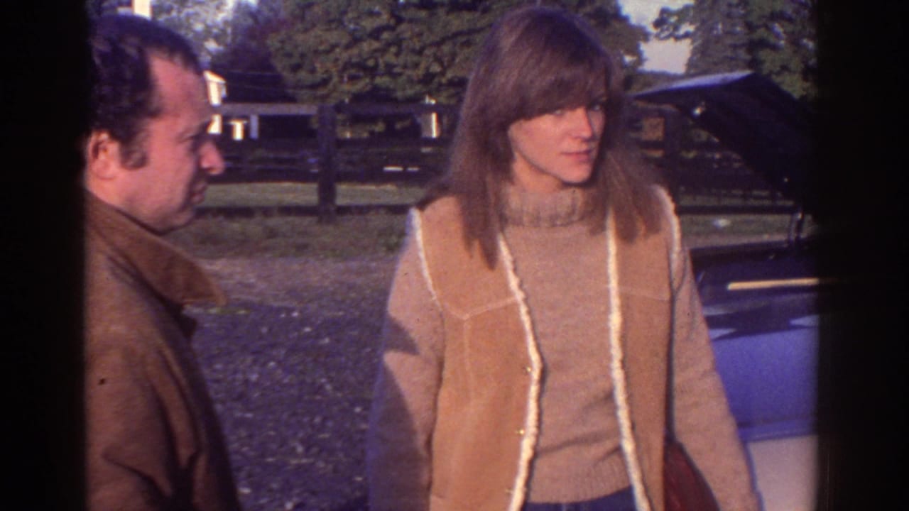 The woman in a tan shearling vest looks toward the car while the man stands partly behind her in Watkins Glen, 1976.