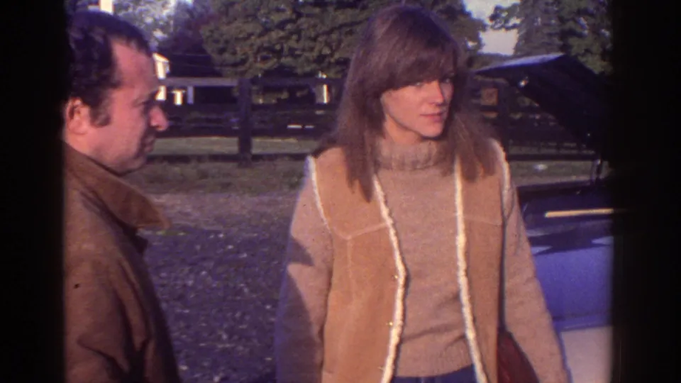 The woman in a tan shearling vest looks toward the car while the man stands partly behind her in Watkins Glen, 1976.