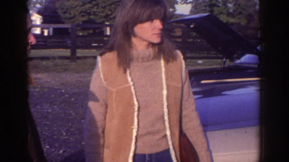 A woman in 1970s clothing stands by a light-colored car with its hood open, with a fence and trees behind her.