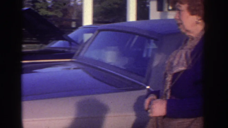 An older woman in a purple jacket stands at the side of the car during the 1976 Watkins Glen home-movie scene.
