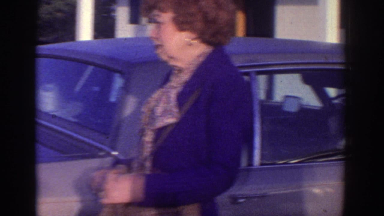 1976 Watkins Glen home-movie frame showing a woman near an older car outside a building with a man partly visible at the side.