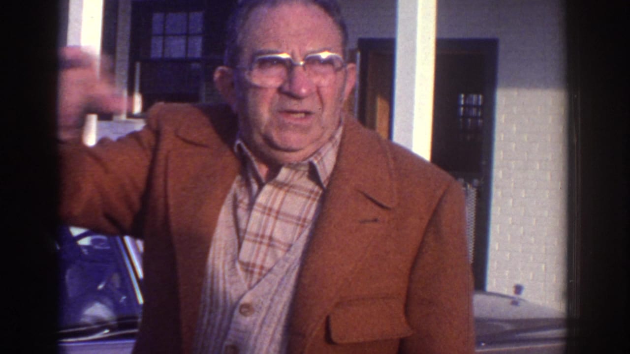 Man in a brown jacket gestures near a parked car outside a building with white pillars.