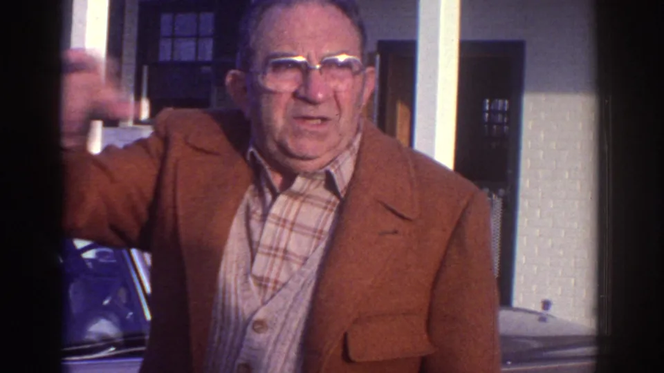1976 Watkins Glen frame of a man in glasses and a brown jacket gesturing beside a parked car near white porch pillars.