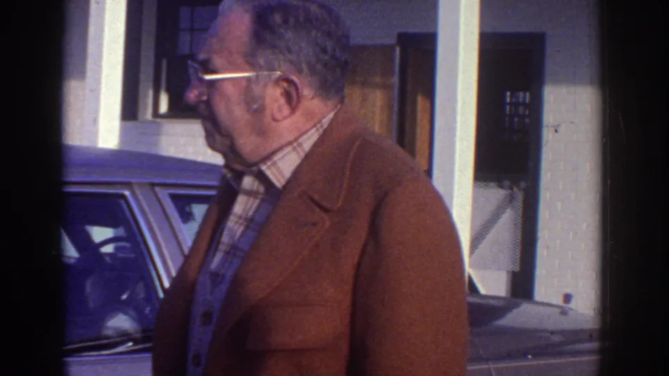 1976 Watkins Glen home-movie frame showing a man in profile near an older car and white-pillared exterior.