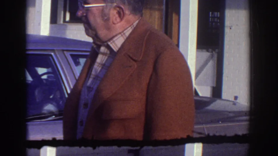 1976 Watkins Glen frame with the man near the parked car, white pillars, and building facade visible in soft 8mm color.