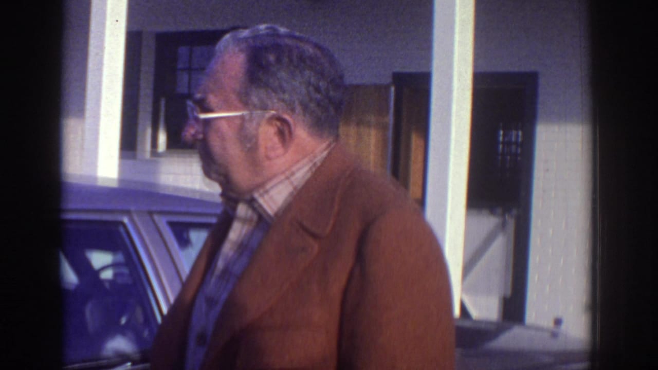 Older man in a brown jacket and plaid shirt stands near a classic car on a Watkins Glen street in 1976 home-movie footage.
