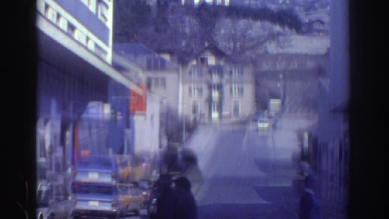 Watkins Glen street view from 1976 home-movie footage, with parked cars, low buildings, and hillside houses beyond.