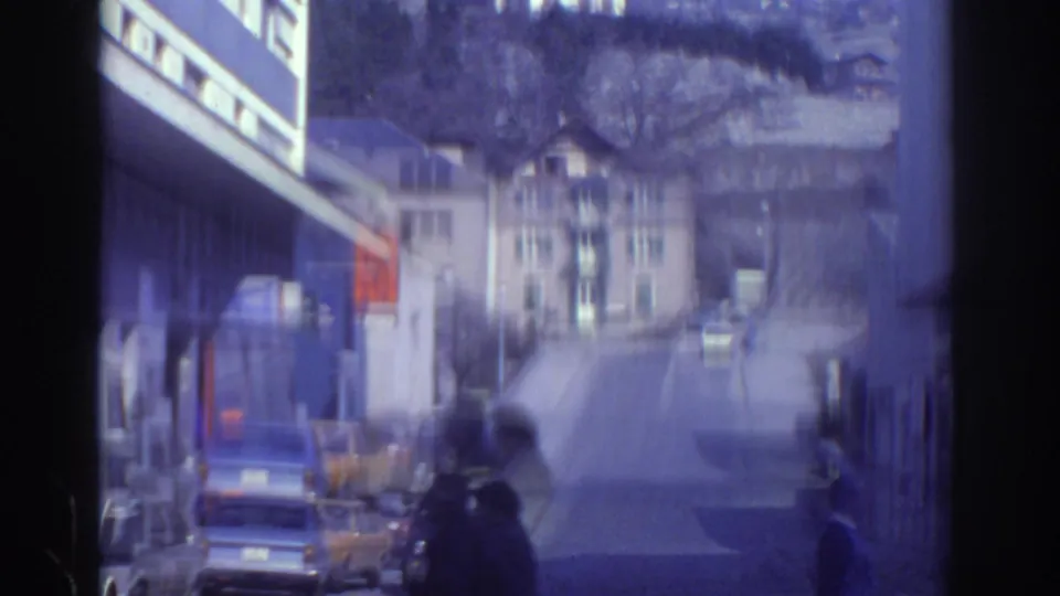 Watkins Glen street view from 1976 home-movie footage, with parked cars, low buildings, and hillside houses beyond.