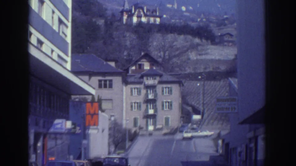 1976 Watkins Glen street scene showing road signs, parked cars, multi-story buildings, and houses rising on the hillside.