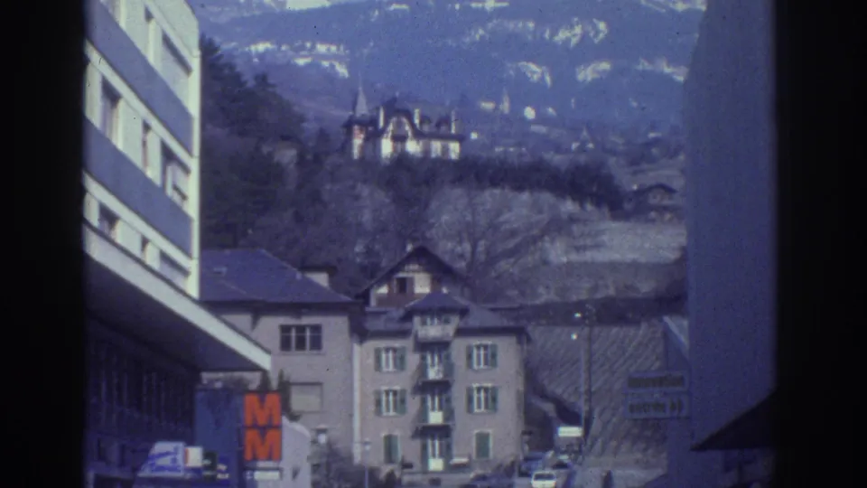 Watkins Glen 1976 home-movie frame with hillside homes, street signs, parked cars, and low-rise buildings in soft 8mm focus.