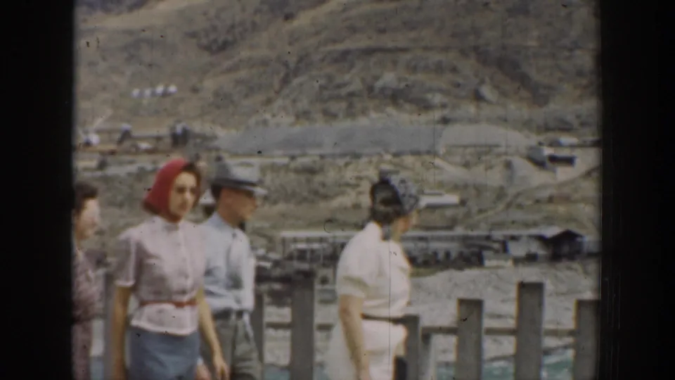 Adult visitors in 1930s clothing stand at a viewing platform overlooking a California dam construction landscape.