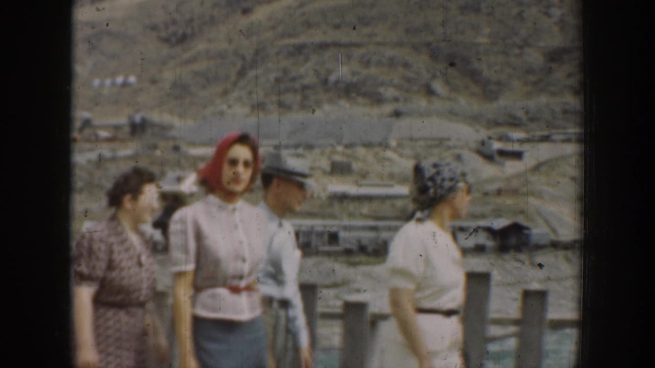 Adults walking by a fence with arid hills and low structures behind them.