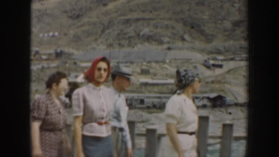 1937 California home-movie frame of adults in period clothing walking beside a fence in a dry rural landscape.