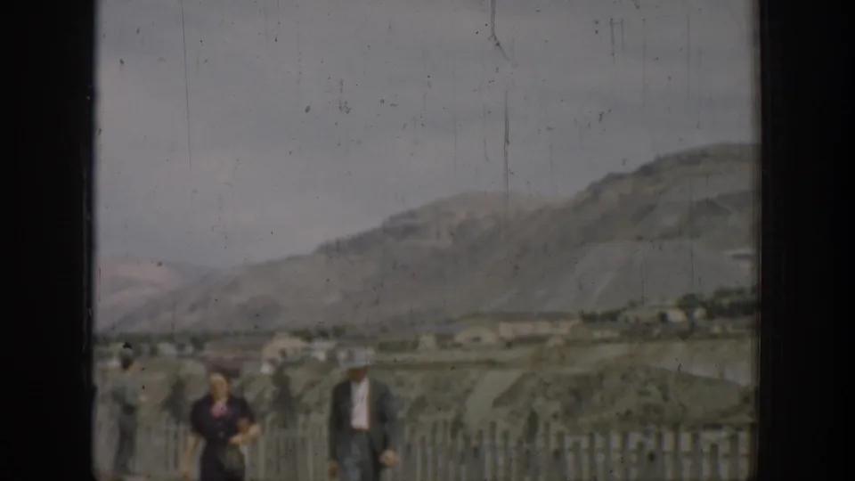 Wide 1937 California home-movie view of adults near a wooden fence below arid hills and an overcast sky.